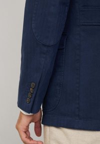 Chaqueta azul marino de tejido texturizado, con solapas de muesca y cuatro botones marrones en el puño. Bolsillo lateral visible en la manga izquierda.