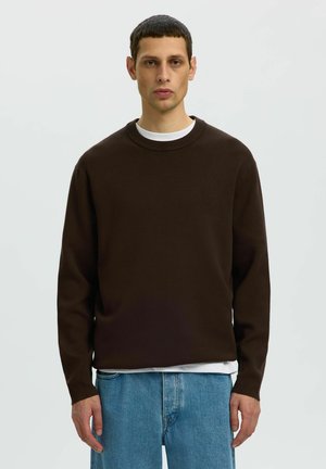 Mann trägt dunkelbraunen Pullover mit Rundhalsausschnitt über weißem Hemd und blauen Jeans, steht vor einem schlichten hellgrauen Hintergrund.