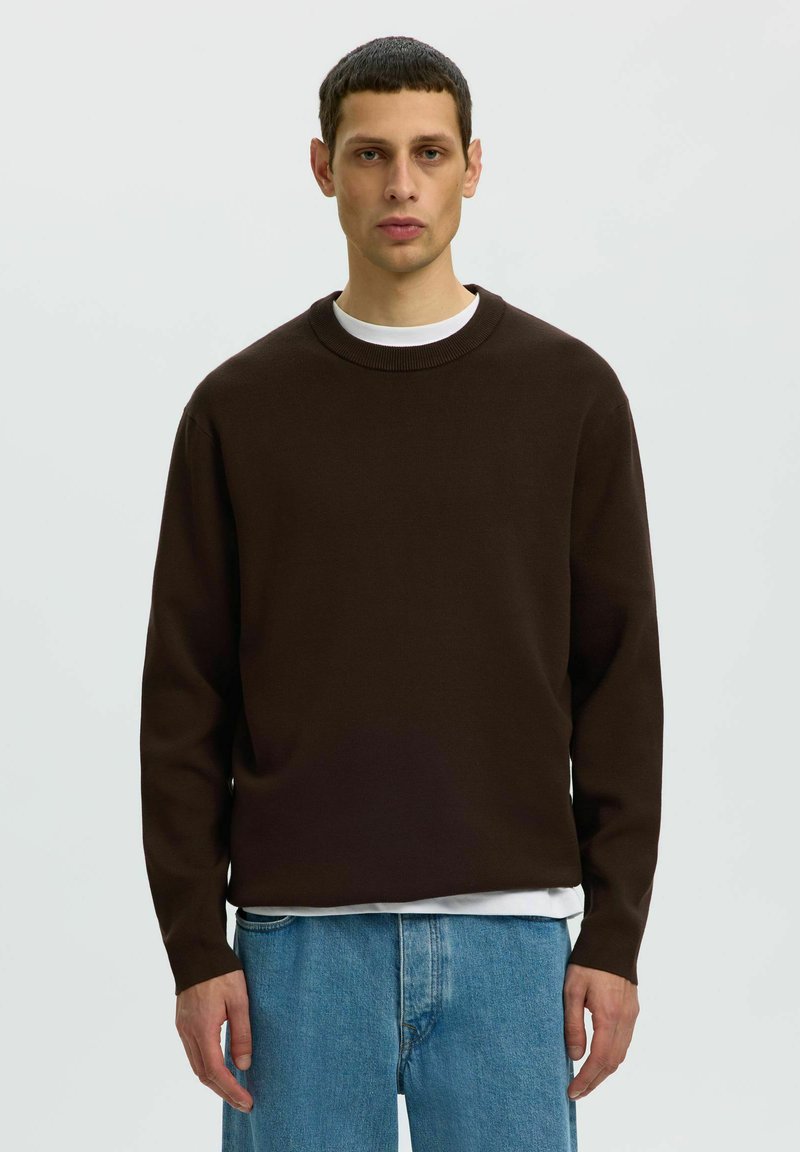 Homme portant un pull marron foncé à col rond par-dessus une chemise blanche et un jean bleu, debout devant un fond uni gris clair.