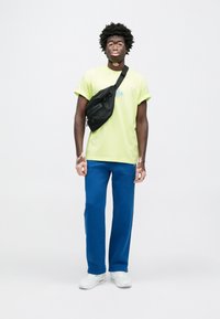 Λαδί πράσινο t-shirt με μπλε λογότυπο, μπλε φόρμα και λευκά αθλητικά παπούτσια. Μαύρη τσάντα χιαστί με φερμουάρ. Ομαλή υφή.