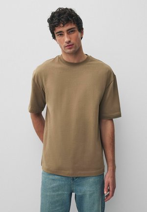 Junger Mann mit lockigem dunklem Haar, der ein locker sitzendes braunes kurzärmeliges T-Shirt und hellblaue Jeans vor einem schlichten hellen Hintergrund trägt.