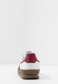 Vit sneakers med en röd läderhäll, strukturerade tygdetaljer och en brun gummisula, med en låg design och rund tå.