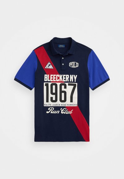 Tengerészeti kék polo ing vörös és kék átlós színblokkokkal. A feliratok között szerepel a "BLEECKER NY 1967" és a "RALPH LAUREN ROAD RUNNERS" fehér színben.