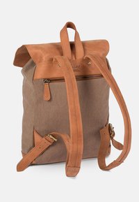 Brown canvas backpack with a tan leather flap, adjustable straps, and a zippered pocket. Features gold-tone hardware and a textured finish.