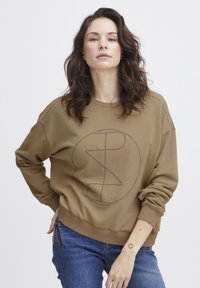 Barna pulóver, amelyen egy nagy, kör alakú grafikai minta látható, szegecsekkel körbevéve. Az anyaga puha, bordás mandzsettákkal és alsó szegéllyel. Kék farmerral párosítva.