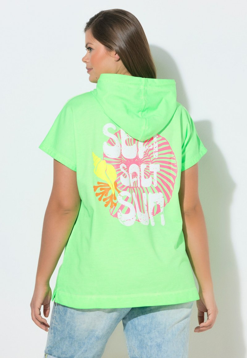 Vrouw met de rug naar de camera, die een felgroene katoenen korte mouw capuchon top draagt met roze en gele schelpenprint en witte tekst "SURF SALT SURF."
