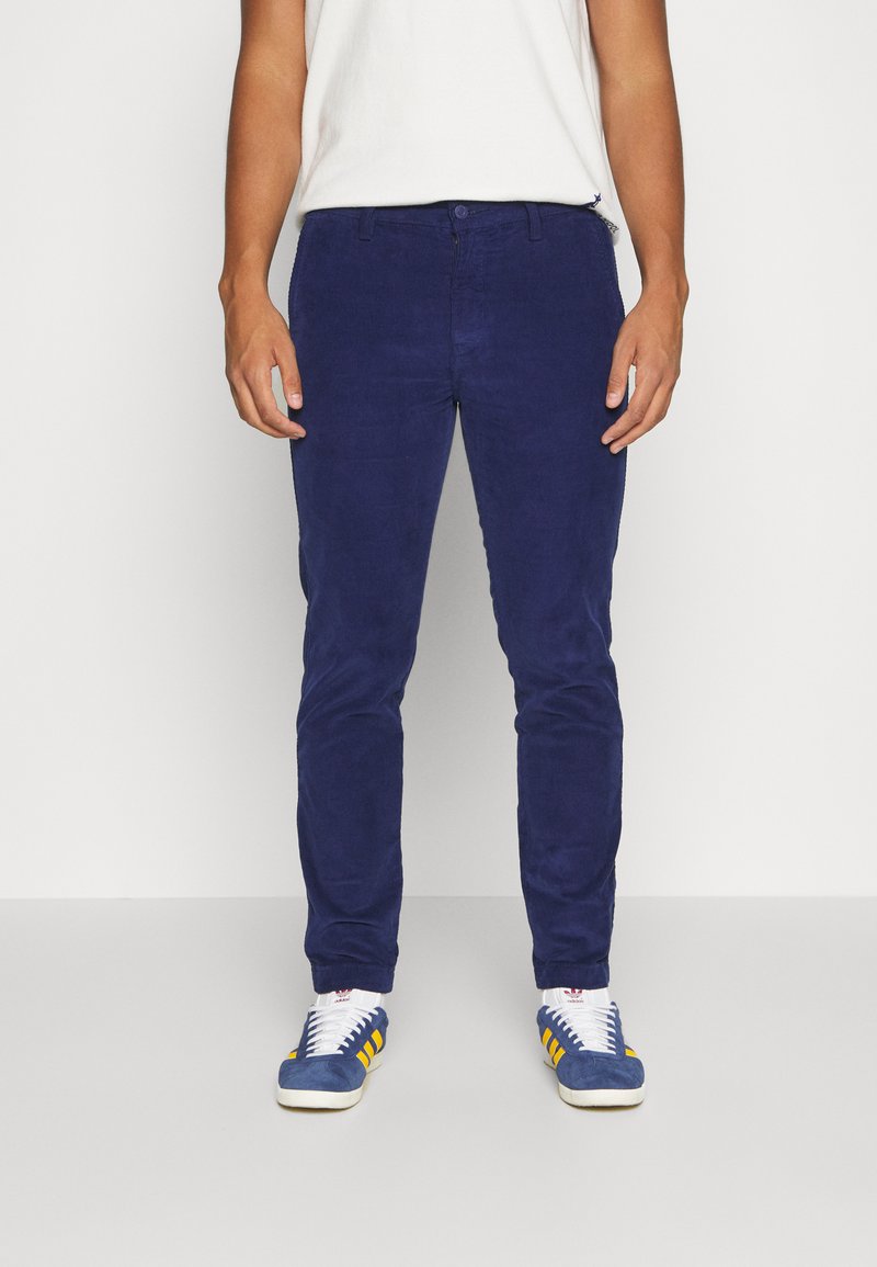 Pantalon en velours côtelé bleu marine avec une coupe slim, tissu texturé, fermeture par bouton et zip, et poches avant, associé à des baskets bleues avec des rayures jaunes.