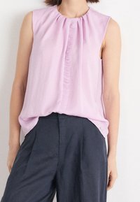 Blusa sin mangas de lavanda con cuello redondo fruncido y detalle de volantes en la parte delantera, combinada con pantalones oscuros de tiro alto.