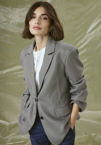Šedý vzorovaný blazer s klopovým límcem, dvěma knoflíky a nastavenými kapsami, nošený přes bílý top a doplněný modrými džíny.