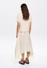 Light cream top with short sleeves and a relaxed fit. Flowing, asymmetrical cream skirt with scalloped edges and decorative stitching. Black sandals.