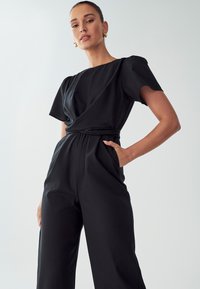 Zwarte jumpsuit met korte mouwen, een wikkeldesign aan de voorkant en zijzakken. Gladde stof met een relaxte pasvorm en een gedefinieerde taille.