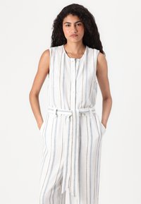 Ermeløs, belte jumpsuit i et lett stoff med vertikale striper i blått og brunt på en hvit bakgrunn, med frontknapper og lommer.