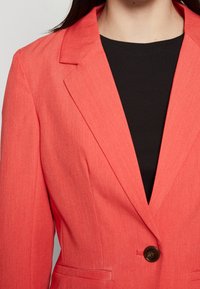 Korallfärgad blazer i en något texturerad tygkvalitet, med enkel knappstängning och slag. Bärs över en svart ribbad topp.