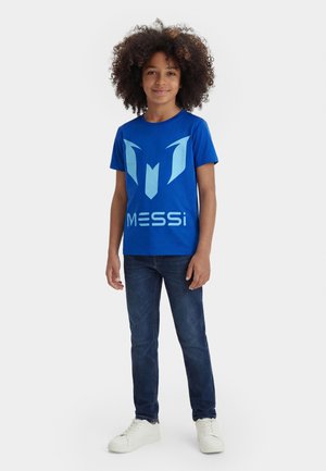 Joven con cabello rizado que lleva una camiseta azul de Messi, vaqueros oscuros y zapatillas blancas, posando frente a un fondo liso.