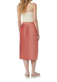 Geplisseerde midi-skirt in een zachte koraalkleur, met een aansluitende tailleband en een vloeiende ontwerp, gecombineerd met lichte sandalen. Glad textuur.