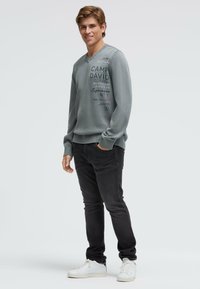 Grauer V-Ausschnitt-Pullover mit bedrucktem Text und Logo, kombiniert mit schwarzer Jeans und weißen Turnschuhen, mit gerippten Bündchen und Saum.