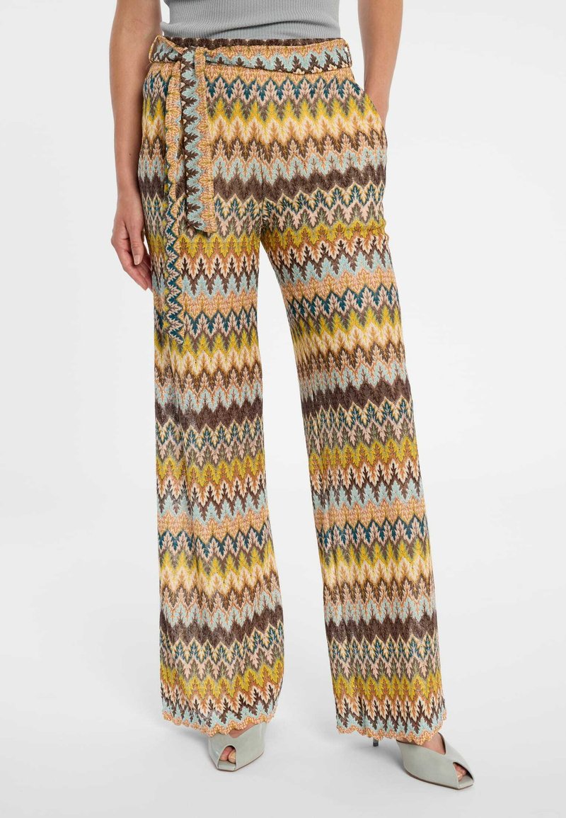 Wijde broek met meerkleurig zigzagpatroon en bijpassende stoffen riem, gedragen met een mouwloze grijze top en lichtgroene open teen hakken.