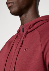 Bordeauxroter Reißverschluss-Hoodie aus strukturiertem Stoff, mit einem kleinen schwarzen Nike-Logo auf der Brust und Kordeln an der Kapuze.