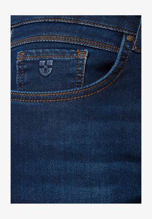 Kieszonka w dżinsach z pomarańczowym przeszyciem i małym haftowanym emblematem na kieszeni na monety, ukazująca teksturę i detale materiału.