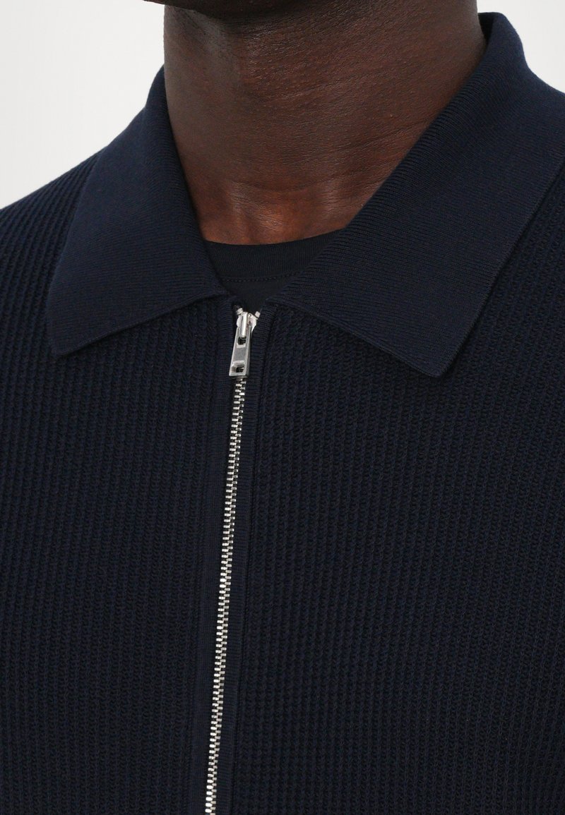 Marineblå strikket cardigan med ribbet tekstur, der har en flad krave og sølvfarvet lynlås lukning fortil.