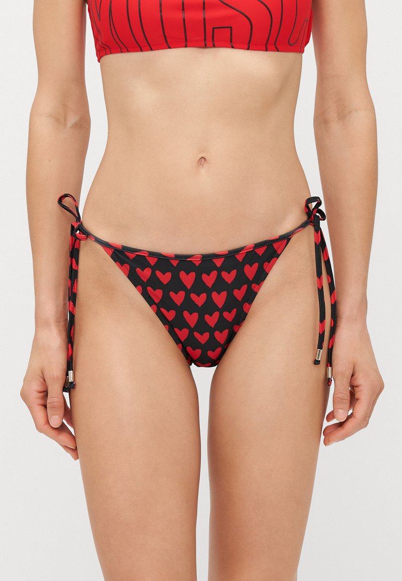 Melns bikinis ar sarkanu sirsniņu rakstu, ar regulējamiem sānu saišu elementiem un metāliskiem akcentiem. Gluda, elastīga auduma.