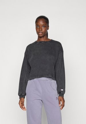 Svart kort sweatshirt i slitt stil, med lange ermer og en liten logo på brystet. Kombinert med lyse lilla joggebukser.