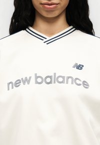 Γυναίκα που φοράει εκρού μπλούζα New Balance με λαιμόκοψη σε σχήμα V με ρίγες σε ναυτικό μπλε και λογότυπο New Balance στο στήθος σε κοντινό πλάνο.