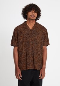 Uomo con capelli ricci che indossa una camicia a maniche corte con stampa astratta marrone e nera, in piedi davanti a uno sfondo neutro.