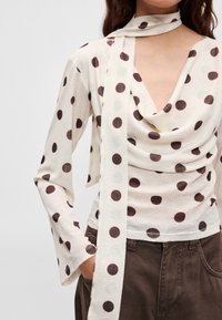 Kremowy top w brązowe groszki, z luźnym dekoltem, długimi rękawami i dopasowanym szalikiem. Materiał wydaje się miękki o delikatnej fakturze.