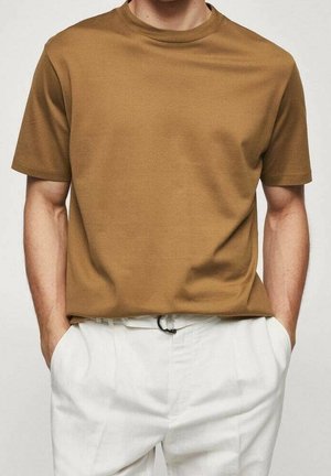 Homme portant un t-shirt marron uni à manches courtes rentré dans un pantalon blanc, les mains dans les poches, sur un fond neutre.