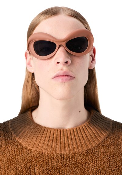 Bruna, stora solglasögon med en tjock, rundad båge och mörka linser. Texturerad, klumpig brun stickad tröja med ribbad krage.