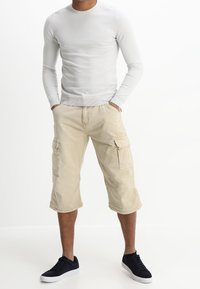 Shorts cargo beiges avec poches latérales, associés à une chemise ajustée à manches longues grise et des baskets noires. Design simple et décontracté.