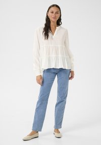 White long-sleeve blouse with a V-neck and tiered ruffle design, paired with light blue jeans and beige slip-on shoes.