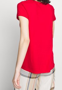 T-shirt rossa a maniche corte con scollo rotondo, realizzata in tessuto leggero, vista di spalle. Abbinata a pantaloni a quadri.