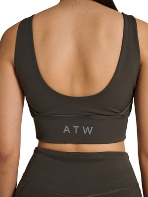 Vrouw draagt een donker olijfgroene sportbeha en hoge leggings, met de rug naar de camera, met de tekst "ATW" op het beha-bandje.