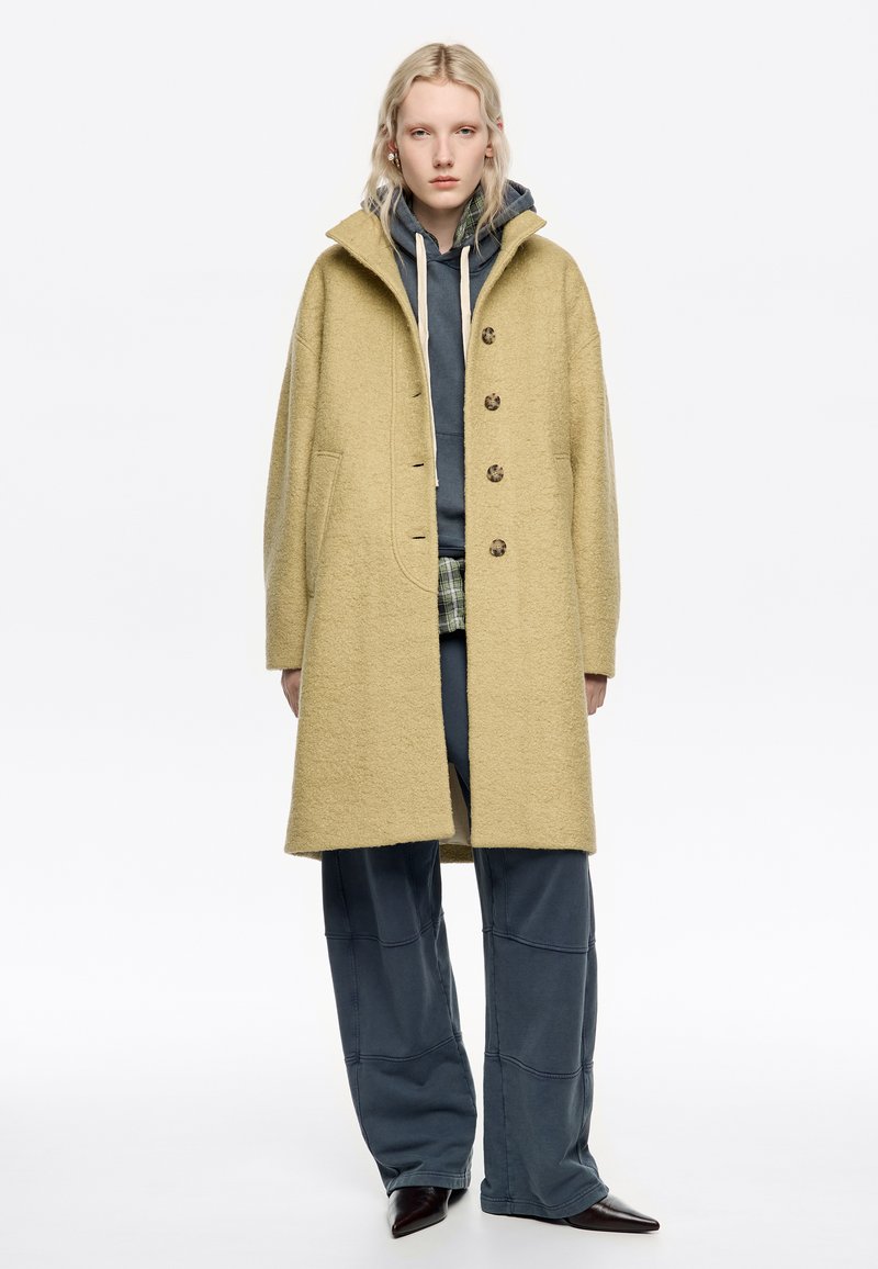 Persona lleva un abrigo largo de lana beige sobre una sudadera oscura, una camisa de cuadros, pantalones oscuros holgados y zapatos oscuros de punta, de pie frente a un fondo blanco.