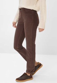 Persona che indossa pantaloni in corduroy marroni, maglione beige e sneakers marroni con suole in gomma, in piedi di fronte a uno sfondo bianco uniforme.