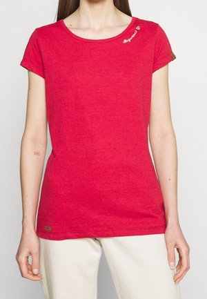 Mujer con camiseta roja de manga corta con texto blanco y un pequeño corazón cerca del cuello, combinada con pantalones de color claro.