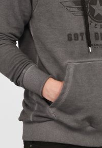 Grå huvtröja med ribbade muddar och nederkant, en framficka och en tryckt logotyp och design över bröstet. Strukturerad bomullsmaterial.