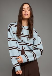 Světle modrý cardigan s hnědými horizontálními pruhy, vyrobený z měkkého materiálu. Má kulatý výstřih a zapínání na knoflíky vpředu.