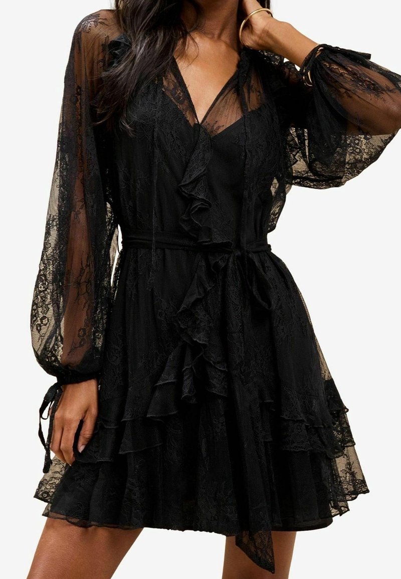 Femme portant une robe en dentelle noire avec des manches ballon transparentes, un devant à volants et une jupe à étages, accessoirisée avec des bracelets.