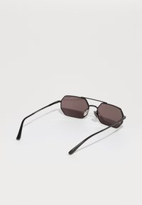 Óculos de sol octogonais com armação em metal preto e lentes roxas escuras. Braços retangulares finos com extremos curvados e acentos prateados na ponte.