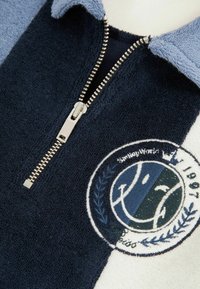 Námornícky modré a svetlomodré fleecový pulóver na zips s terry textúrou; obsahuje vyšívané logo v tvare kruhu so usmievavou tvárou a vavrínovým dizajnom.