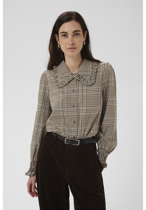 Vrouw met donker haar draagt een beige en zwart geruit blouse met een gerimpelde kraag en manchetten, gecombineerd met een zwarte broek en een riem.