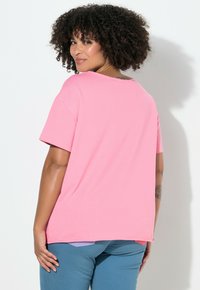 Růžové tričko s krátkým rukávem a kulatým výstřihem, volný střih a jemná textura. Výrazný zaoblený lem, v kombinaci s modrými kalhotami a minimalistickými doplňky.