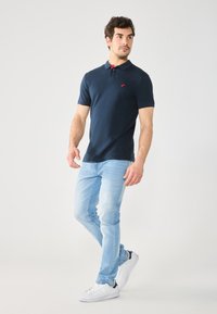 Polo shirt bleu marine avec une patte de boutonnage à trois boutons et un logo d'accent rouge, associé à un jean bleu clair et des baskets blanches.