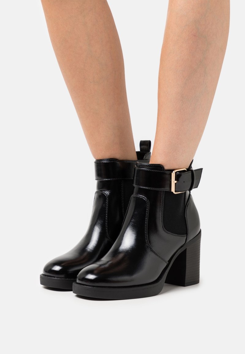 Botas de tobillo de cuero negro brillante con tacones gruesos y hebillas laterales, usadas sobre piernas desnudas contra un fondo blanco.