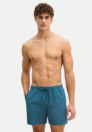 Hombre sin camiseta con cabello rizado, vestido con pantalones cortos de baño color verde azulado, de pie con una mano en el bolsillo contra un fondo liso.
