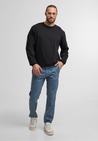 Zwarte longsleeve sweatshirt, blauwe broek, beige sneakers. Duidelijke montuurbril, gladde texturen, relaxte pasvorm, casual uitstraling.