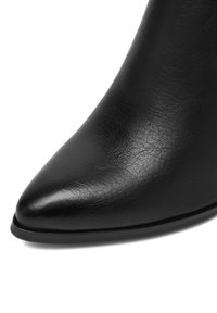 Fekete bőr, hegyes orrú cipő közeli felvétele, sima, enyhén texturált felülettel és vékony fekete talppal, fehér háttér előtt.